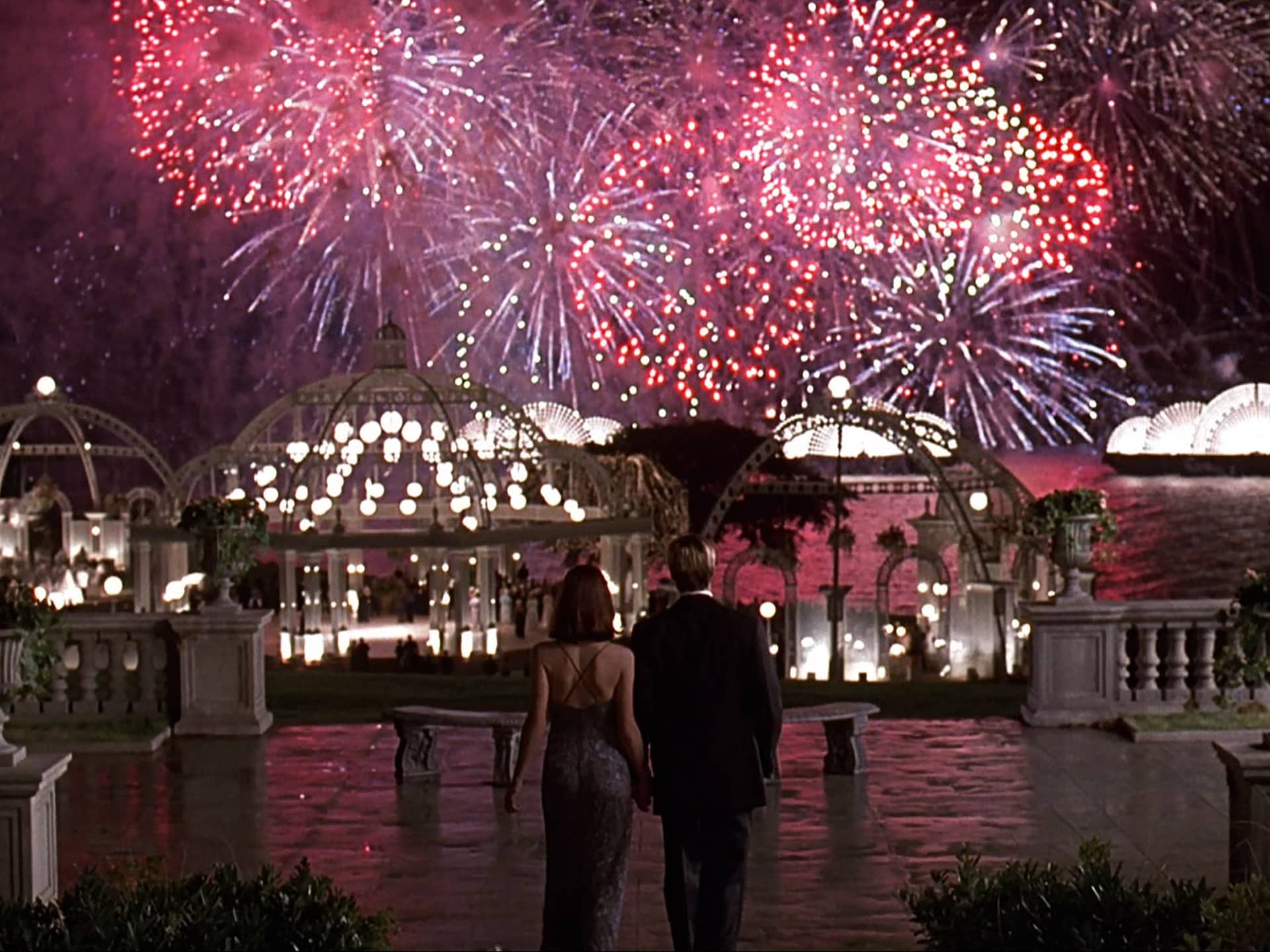A scene from the movie Meet Joe Black, showing Joe Black and Susan Parrish from behind as they hold hands and watch a grand fireworks show over a lavishly lit outdoor party estate.