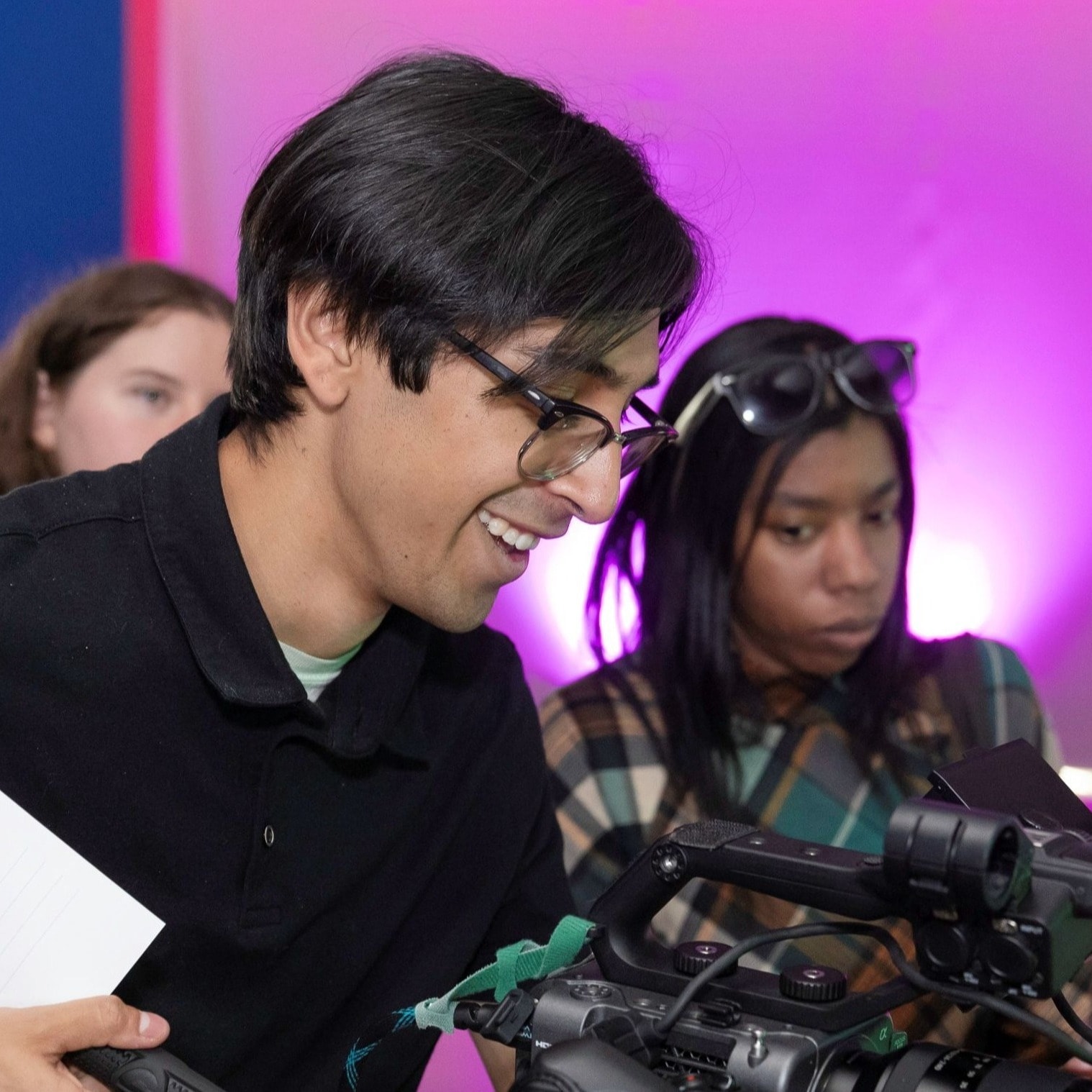 A group of young adults gathered around a professional video camera, focusing on the screen. A young man wearing glasses and a black polo shirt smiles as he looks at the camera setup. The background features vibrant pink and purple studio lighting.