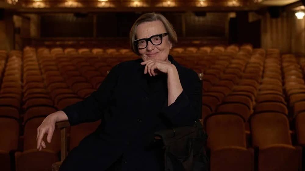 Agnieszka-Holland A portrait of filmmaker Agnieszka Holland seated in an empty theater, resting her chin on her hand and smiling at the camera, surrounded by rows of red velvet seats.