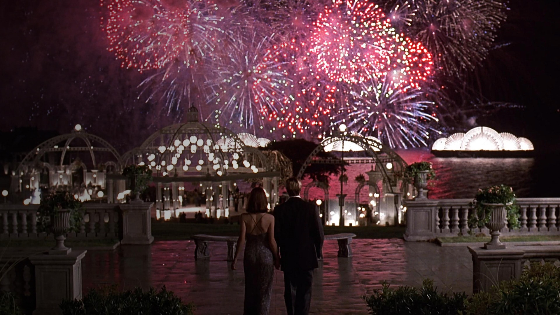 A scene from the movie Meet Joe Black, showing Joe Black and Susan Parrish from behind as they hold hands and watch a grand fireworks show over a lavishly lit outdoor party estate.
